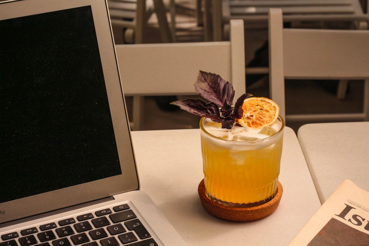 A refreshing citrus cocktail garnished with basil beside a laptop and newspaper, perfect for a casual work setting.